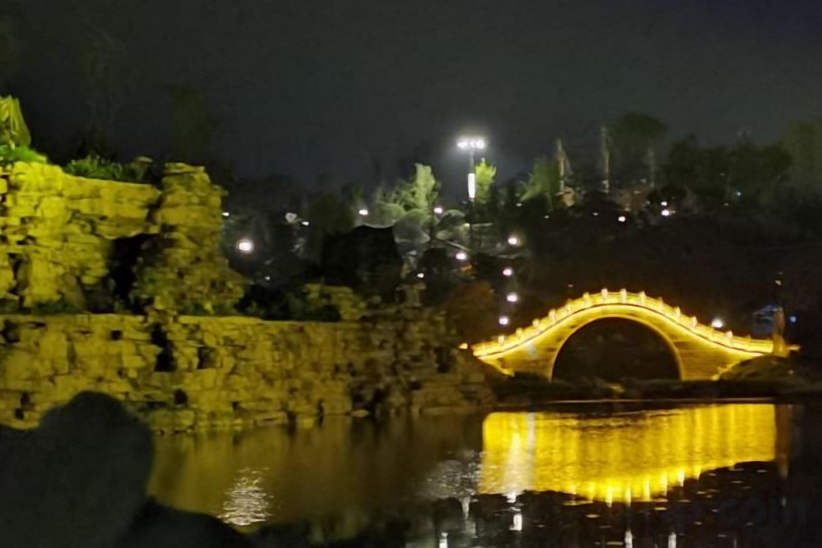 A stone bridge with a reflective surface, illuminated by lights, and a body of water in the foreground.