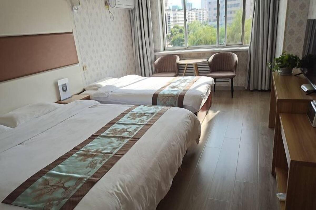Ein Hotelzimmer mit zwei Betten, einem kleinen Tisch, Stühlen, einem Fernseher und einem Fenster mit Blick auf Gebäude.