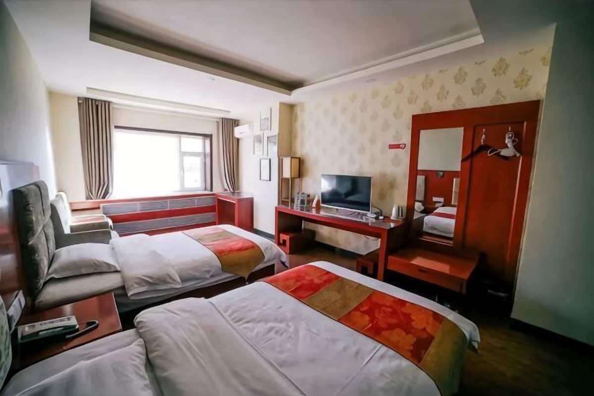 Ein Hotelzimmer mit zwei Betten, einem Schreibtisch mit Fernseher, einem Sessel und einem Fenster mit Vorhängen.