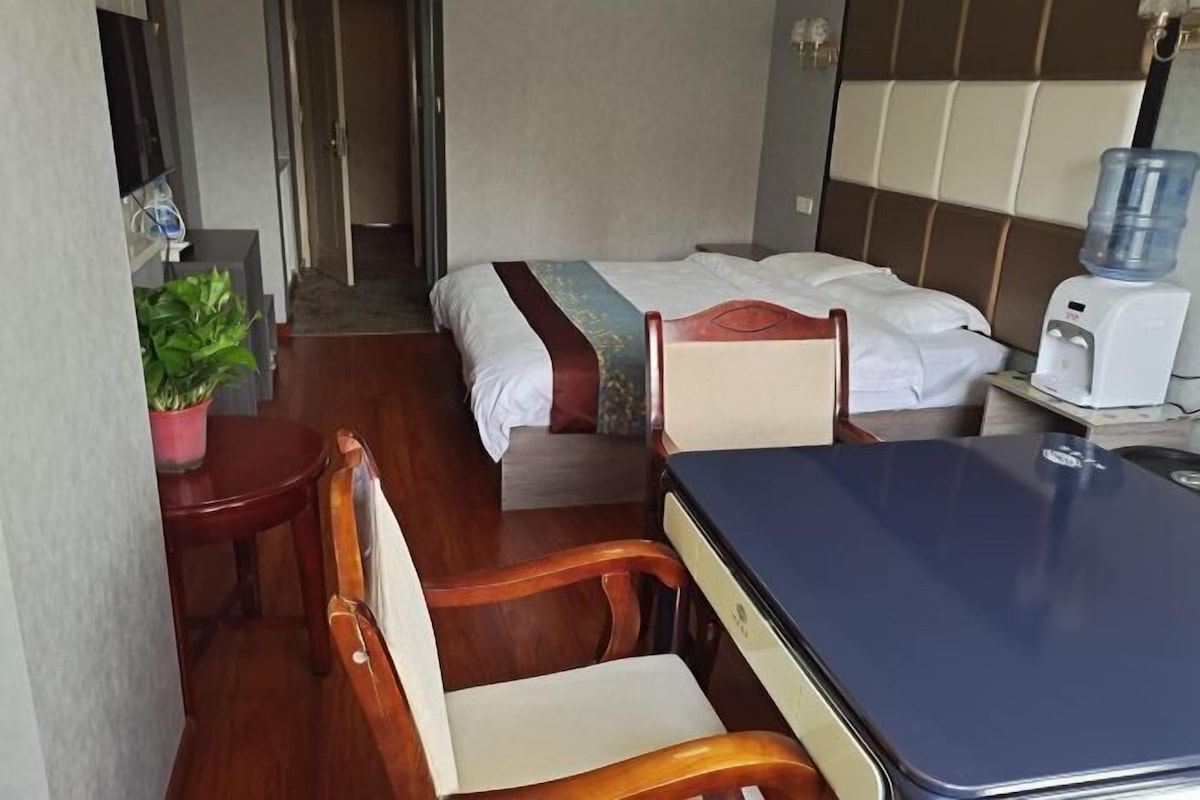 Pieni hotellihuone, jossa on sänky, pieni pöytä, tuoli, vesiautomaatti ja ruukussa oleva kasvi.