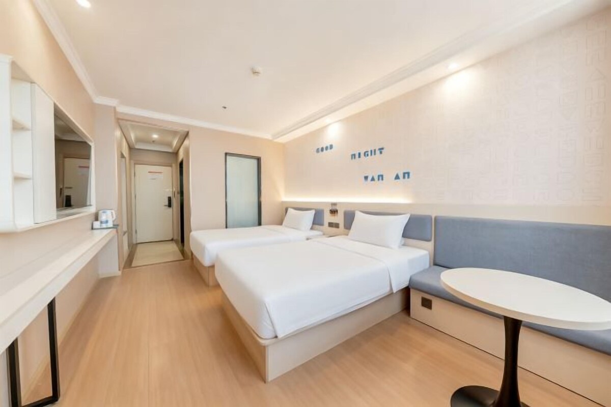 Ein Hotelzimmer mit zwei Betten, einem kleinen Tisch, einer Bank und einem an der Wand befestigten Fernseher.