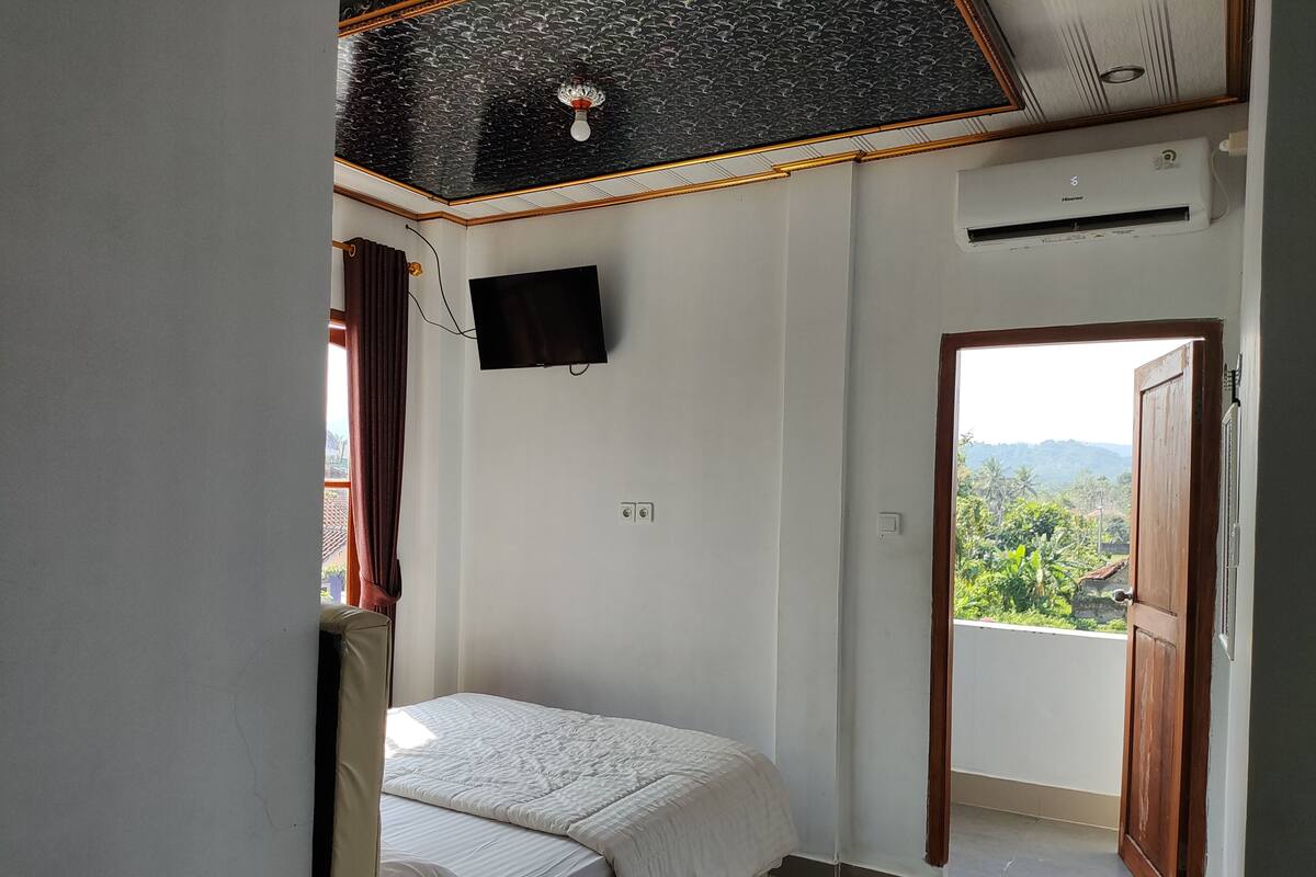 Een slaapkamer met een bed, een wandtelevisie, een airconditioner en een raam met een geopende deur naar een balkon met uitzicht op groen en bergen.