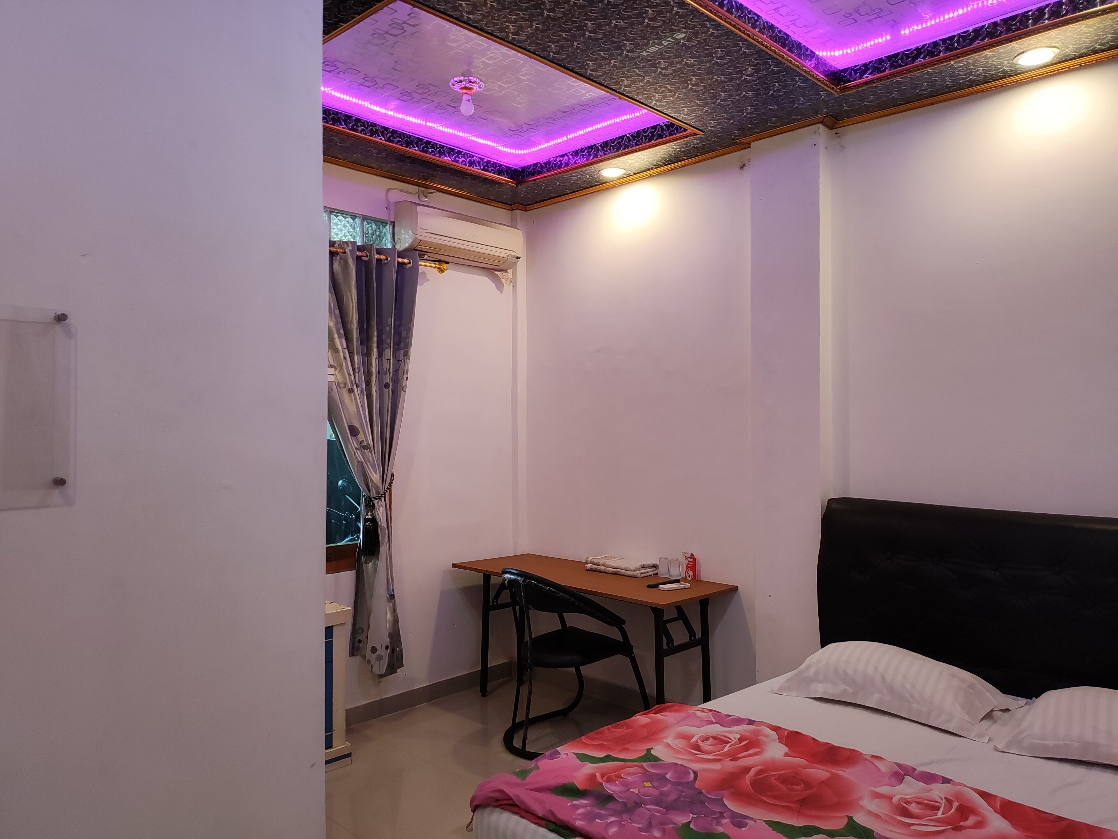 A bedroom with a bed, a desk, and a chair. There is a window with curtains, and the ceiling has decorative lighting.