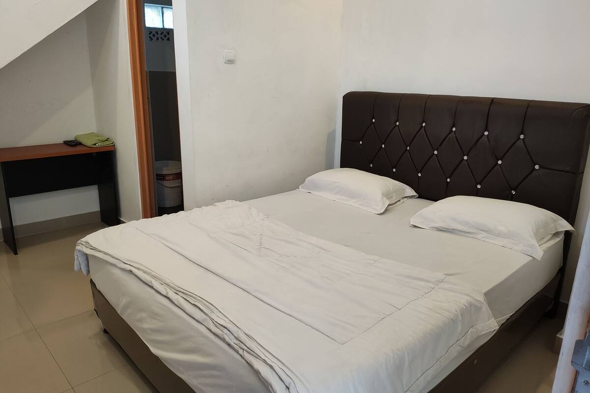 Een net opgemaakt bed met een donker hoofdbord en witte beddengoed in een kamer met een klein tafeltje en een prullenbak.