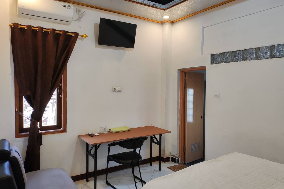 Een kamer met een bed, een bureau met stoel, een aan de muur bevestigde televisie en een raam met gordijnen.