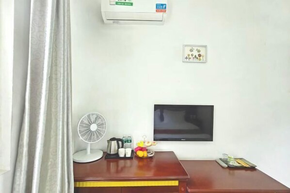 A hotel room with a wooden desk, a flat-screen TV, a standing fan, a small decorative figure, a wall-mounted air conditioner, and a framed picture. - Yunjin Hotel (Yangbi)
