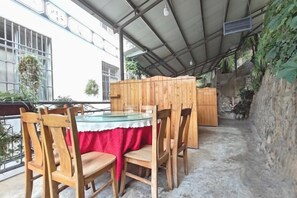 Une terrasse extérieure couverte, avec une table et des chaises, un meuble en bois et une vue sur un bâtiment doté de fenêtres et de plantes.