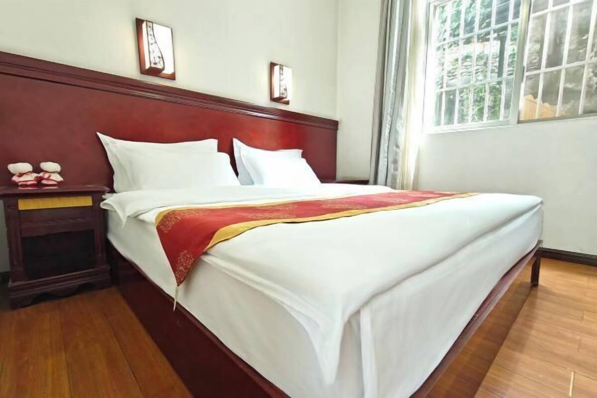 A neatly made bed with white linens and a red decorative throw, a wooden nightstand with a lamp, and a window with a view of greenery.