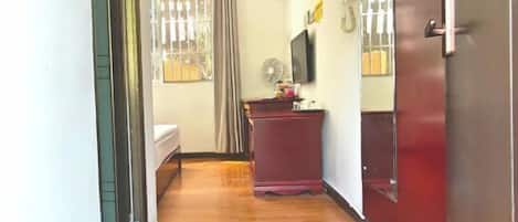 A hallway with a bed, a desk, a fan, and a door.