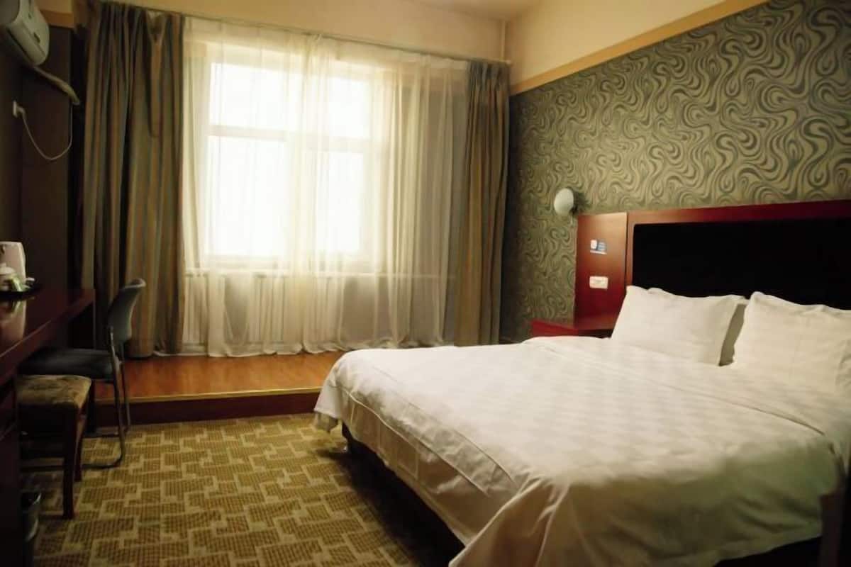 A hotel room with a bed, a desk with a chair, a window with curtains, and a patterned wall.