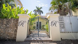 A gated entrance with a stone wall and a metal gate, surrounded by lush greenery and palm trees. - Room-2br Caribbean Condo In Cabarete Cen (Sosúa)