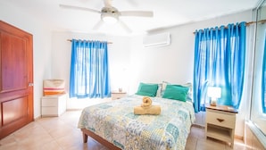 A bedroom with a bed, a nightstand, a lamp, a ceiling fan, and blue curtains. - Room-2br Caribbean Condo In Cabarete Cen (Sosúa)