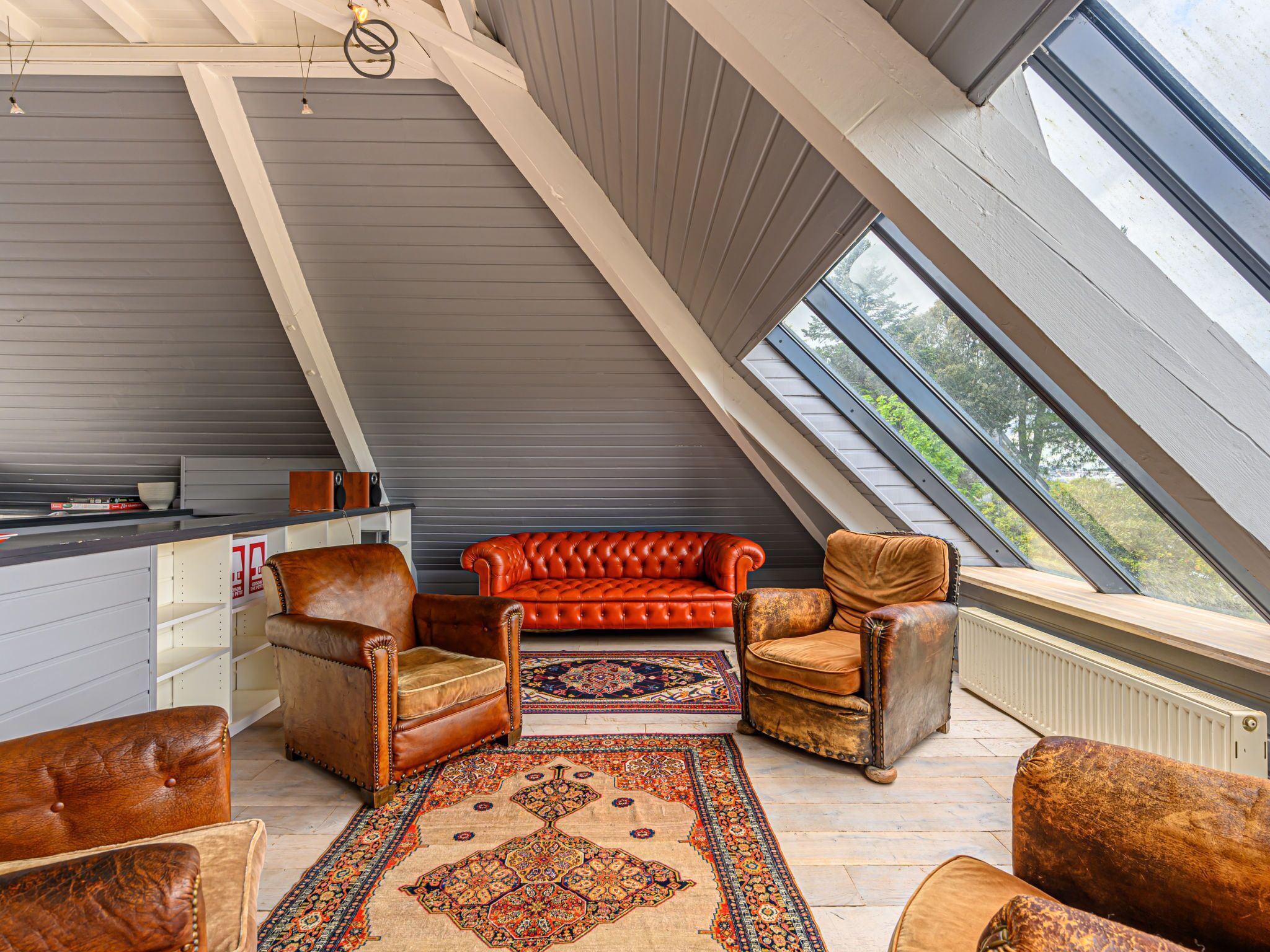 Une pièce de style « loft », avec un plafond haut, un canapé en cuir rouge, des fauteuils en cuir, un tapis à motifs et une vue à travers une lucarne.