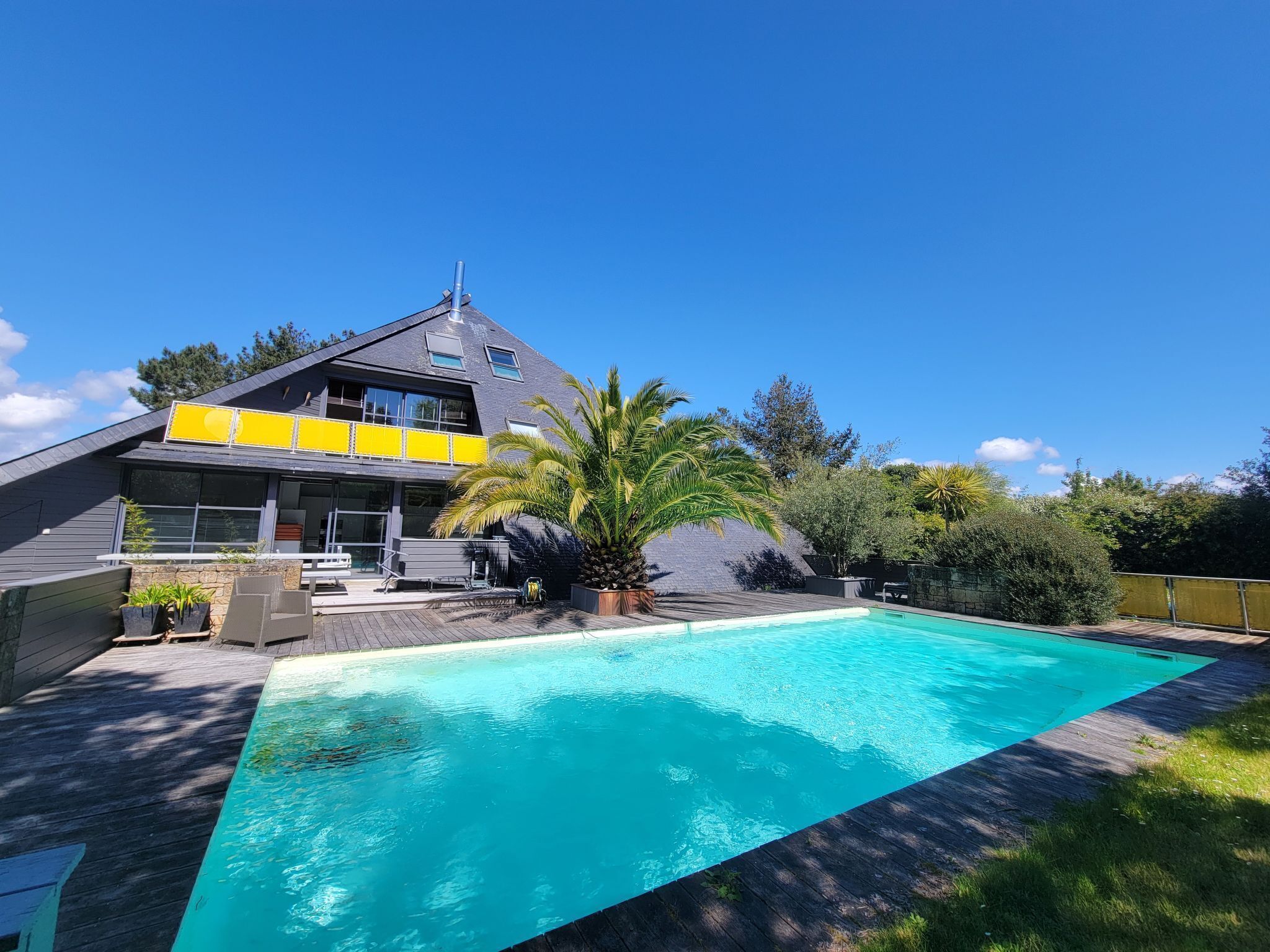 A modern two-story house with a swimming pool, surrounded by greenery and a clear blue sky.