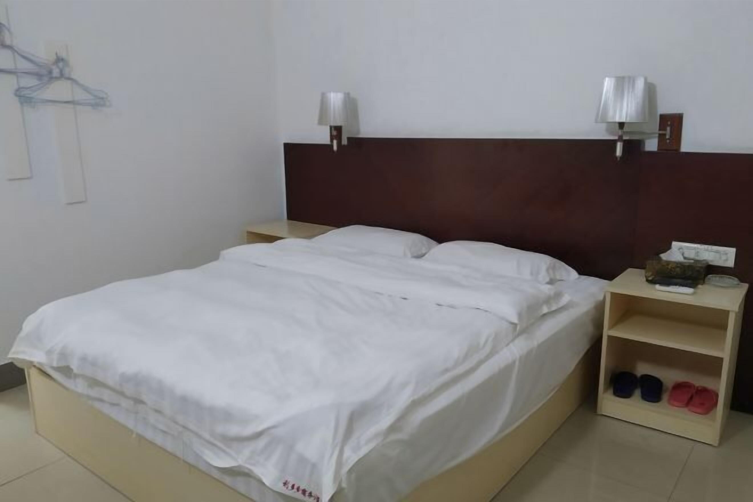 Una cama bien hecha con sábanas blancas, dos lámparas de noche, una mesita de noche con lámpara y un zapatero con tres pares de zapatos.