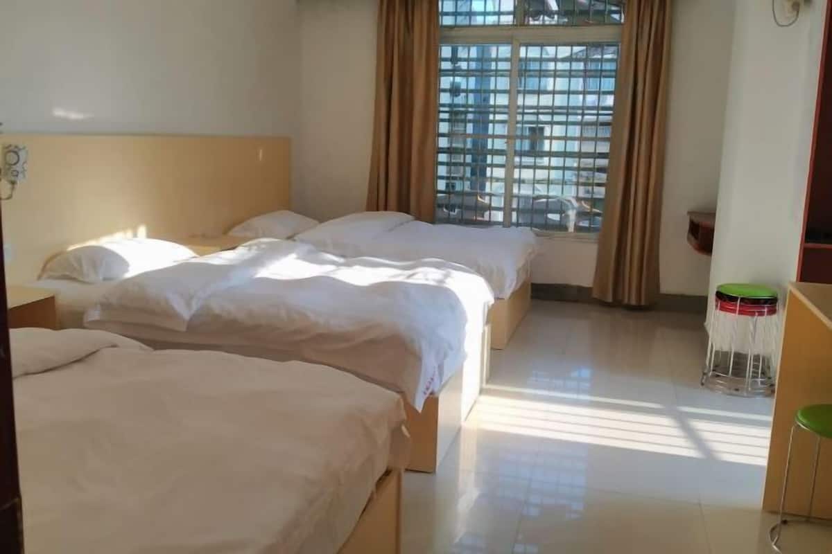 Hotellihuone, jossa on kolme sänkyä, ikkuna, jossa on verhot, pieni jakkara ja laattalattia.