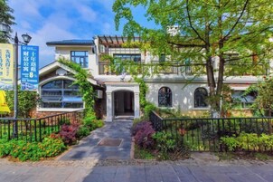 Um edifício de dois andares com estilo arquitetônico tradicional, apresentando uma entrada central com arco decorativo, cercado por vegetação exuberante e um caminho pavimentado que leva à entrada.