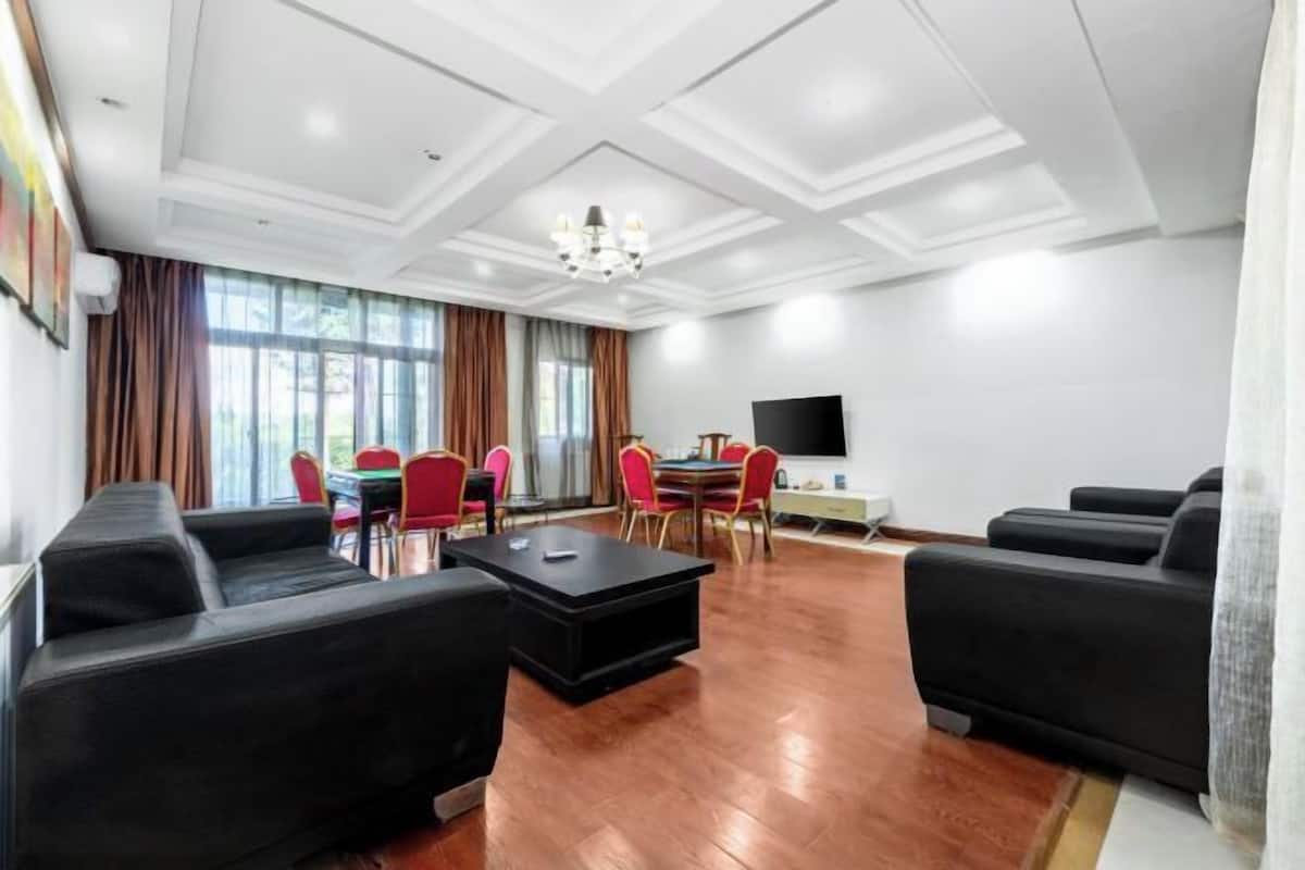 A spacious living room with a large flat-screen TV, black sofas, a dining table with red chairs, and a chandelier.