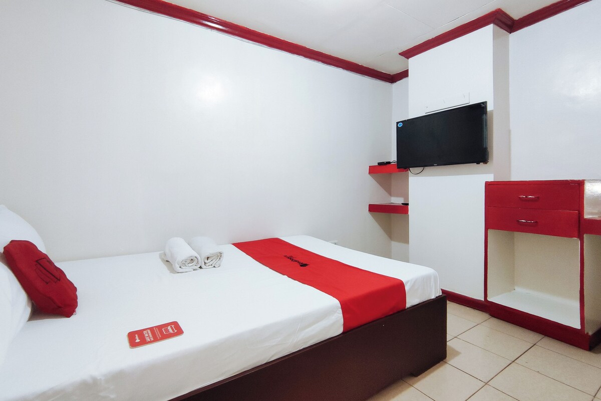 Una habitación de hotel con una cama, un televisor montado en la pared, una decoración en tonos rojo y blanco, y un mueble de almacenamiento rojo.