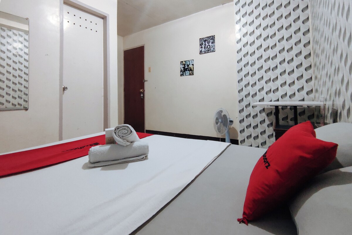 Habitació d'hotel amb un llit, una almoinada vermella, una tovallola, un ventilador i una porta.