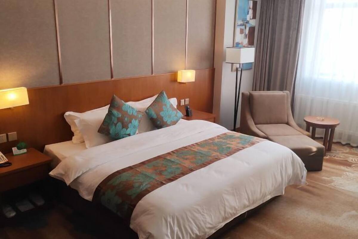 A hotel room with a large bed, a chair, a small table, and a bedside lamp.