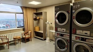 A laundry room with two stacked front-loading washing machines, a drying rack, a chair, and a window with a view of buildings. - Dalian yuanfeng hotel (Dalian)