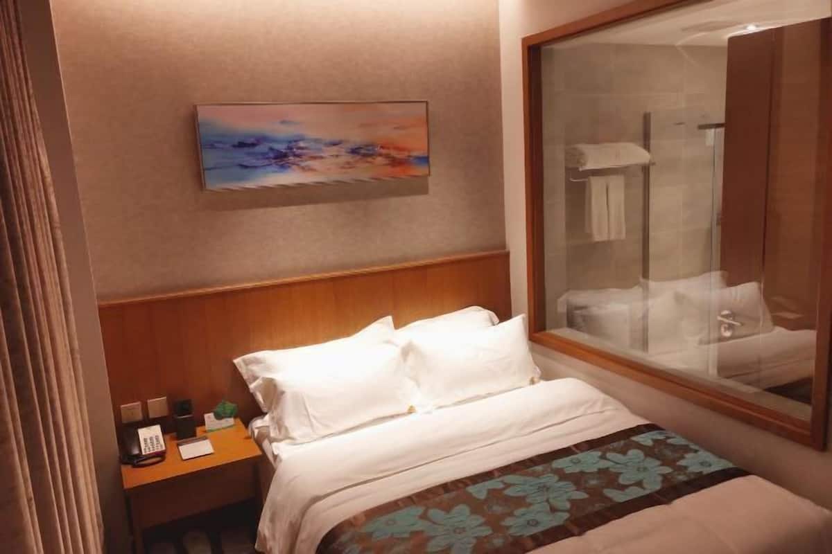 A hotel room with a bed, a bedside table with a phone and a notepad, a painting on the wall, and a large mirror reflecting a bathroom.
