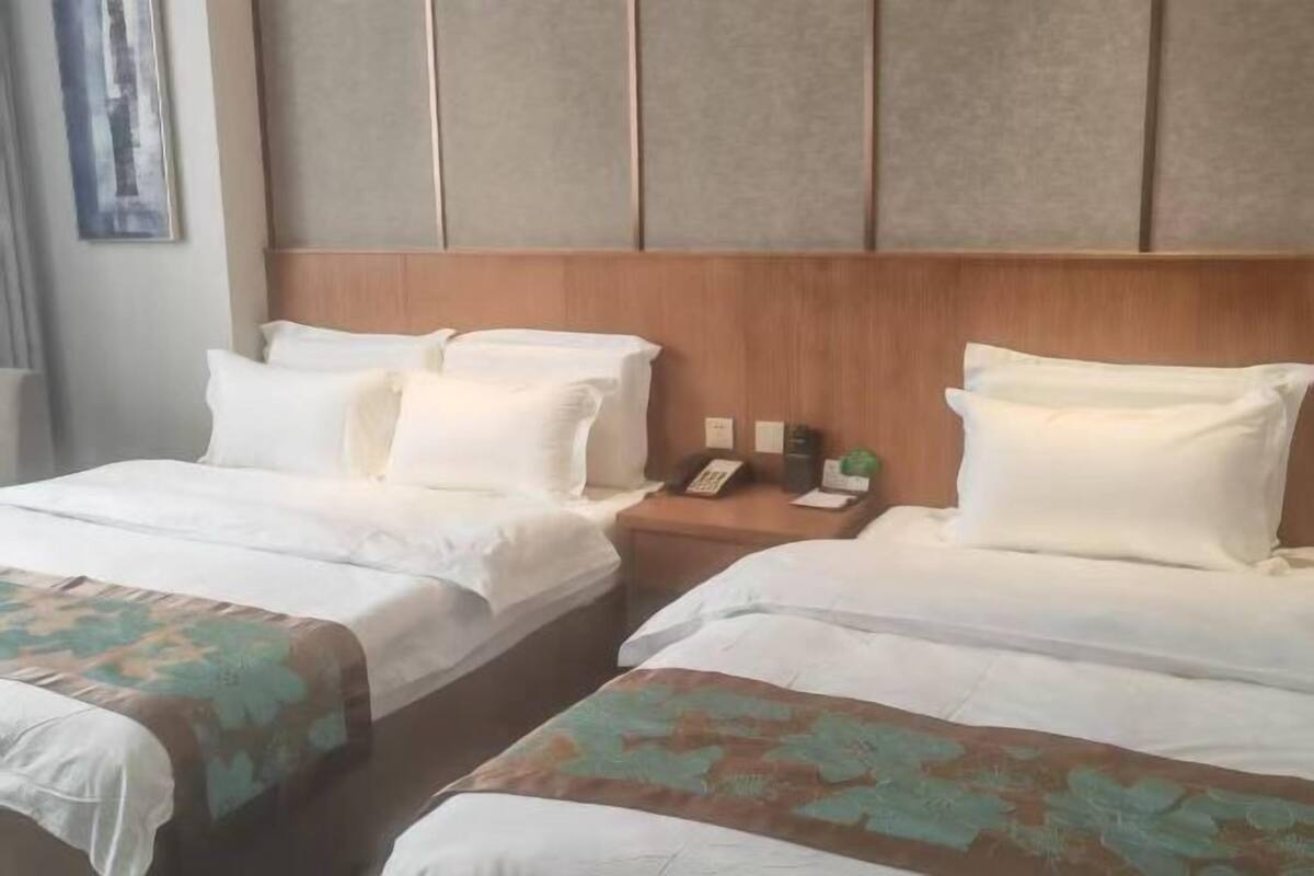 A hotel room with two beds, a wooden headboard, a nightstand with a phone and a plant, and a framed picture on the wall.