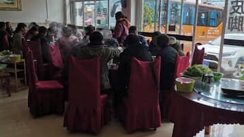 Un grupo de personas sentadas en mesas redondas con manteles rojos, en una sala con ventanas amplias y vista a la calle.