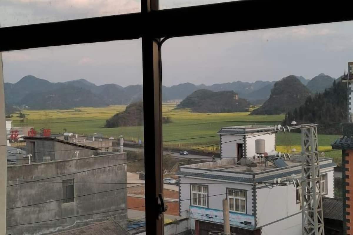 A view through a window of a building with a white exterior, a metal structure on the right, and a landscape with mountains and fields in the background.