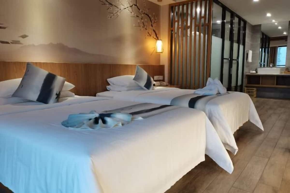 Habitación de hotel con dos camas, cabecero de madera, lámpara de pared y puerta de vidrio corredera, además de baño con lavamanos y espejo.