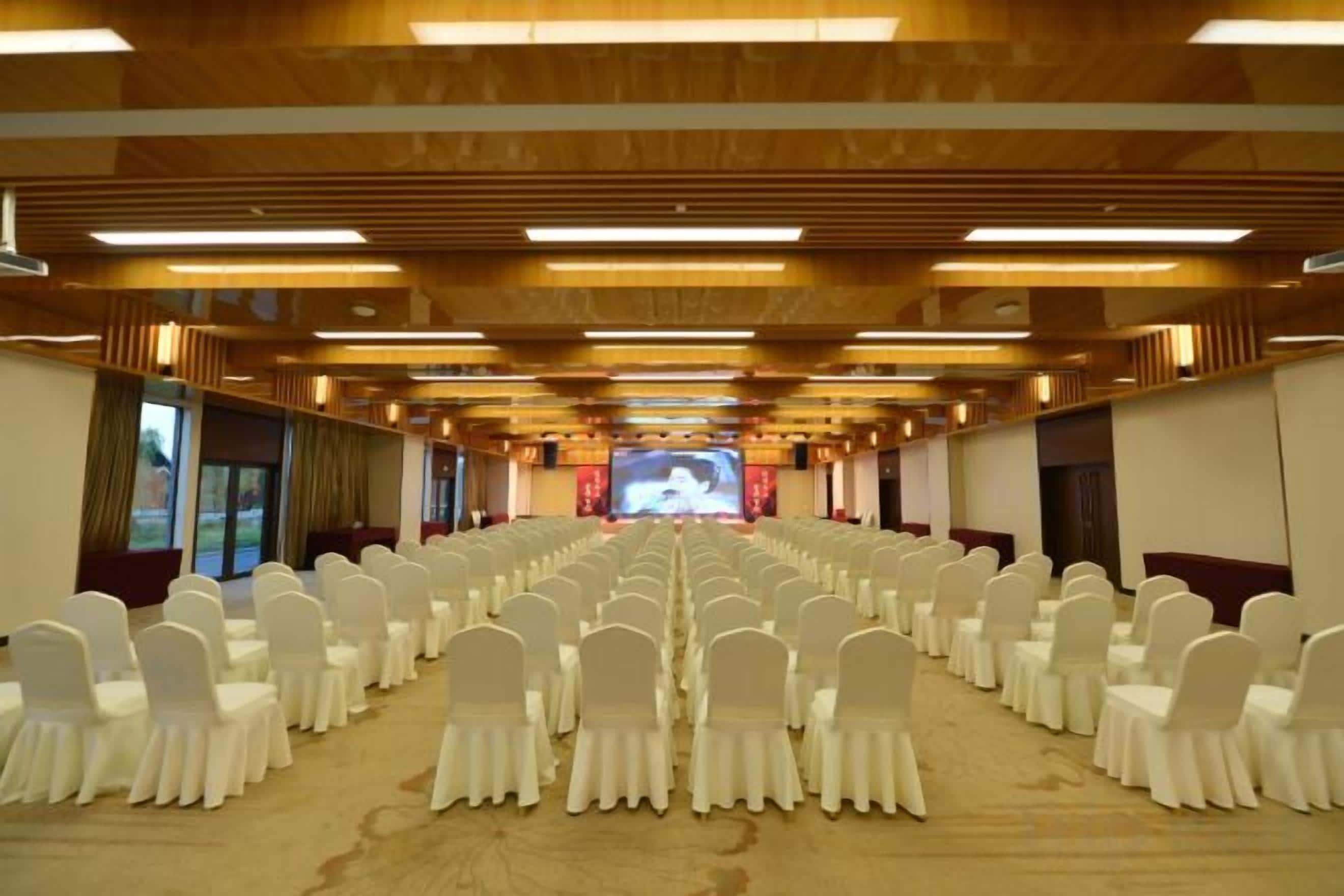 A conference room with rows of white chairs, a large screen at the front, and wooden paneling on the ceiling.