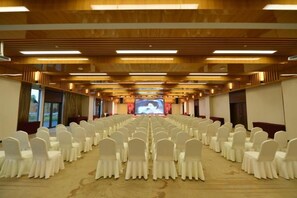 Een vergaderzaal met rijen witte stoelen, een groot scherm aan de voorkant en houten lambrisering aan het plafond.