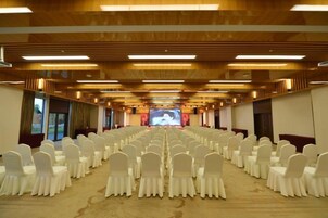Un salón de conferencias con filas de sillas blancas, una pantalla grande al frente y paneles de madera en el techo.