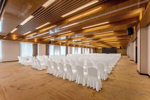 Un amplio salón de conferencias con filas de sillas blancas, paneles de madera en el techo y suelo alfombrado.