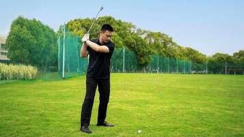A person in black attire on a grassy field, holding a golf club, with trees and a fence in the background.
