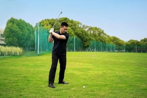 Una persona vestida de negro en un campo de césped, sosteniendo un palo de golf, con árboles y una cerca al fondo.