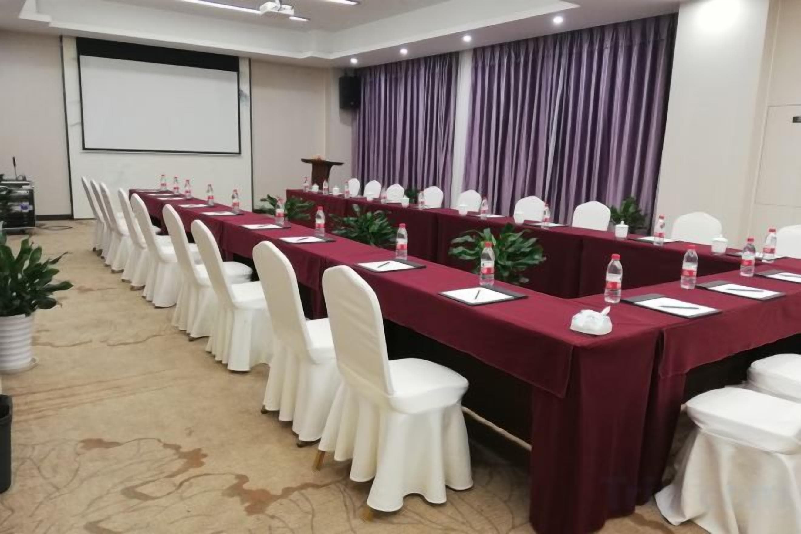 Un salón de conferencias con una larga mesa, sillas blancas y una pantalla de proyector.