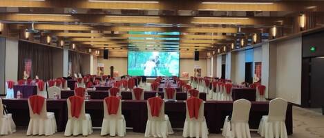 Un salón de conferencias con filas de mesas y sillas, una pantalla grande al frente y una zona escenario.