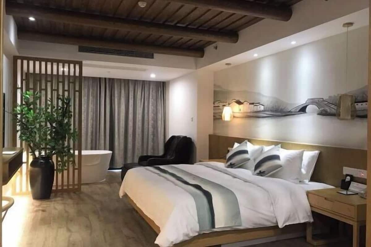 Una habitación de hotel moderna con techo de madera, una cama grande con ropa de cama blanca y gris, un escritorio con una silla, una planta en maceta y una obra de arte en la pared de un puente.