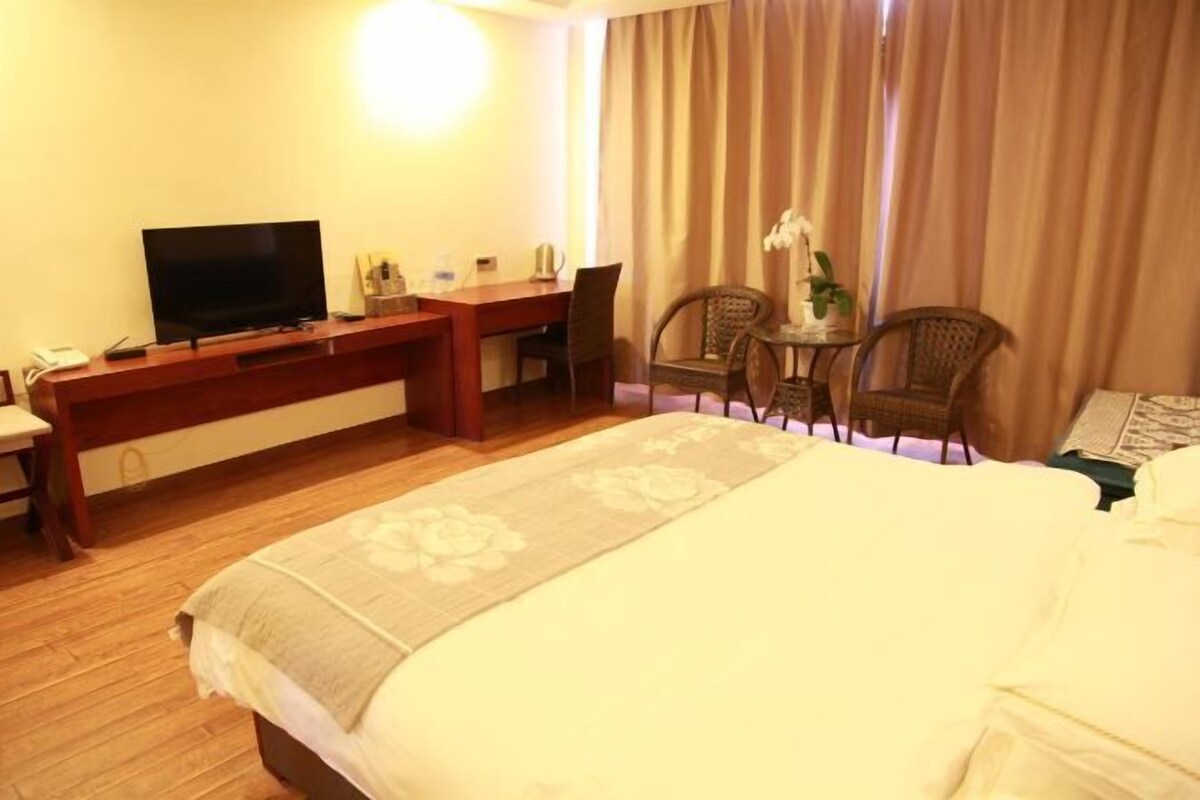 A hotel room with a bed, a desk with a TV, a chair, a small table with a plant, and a window with curtains.