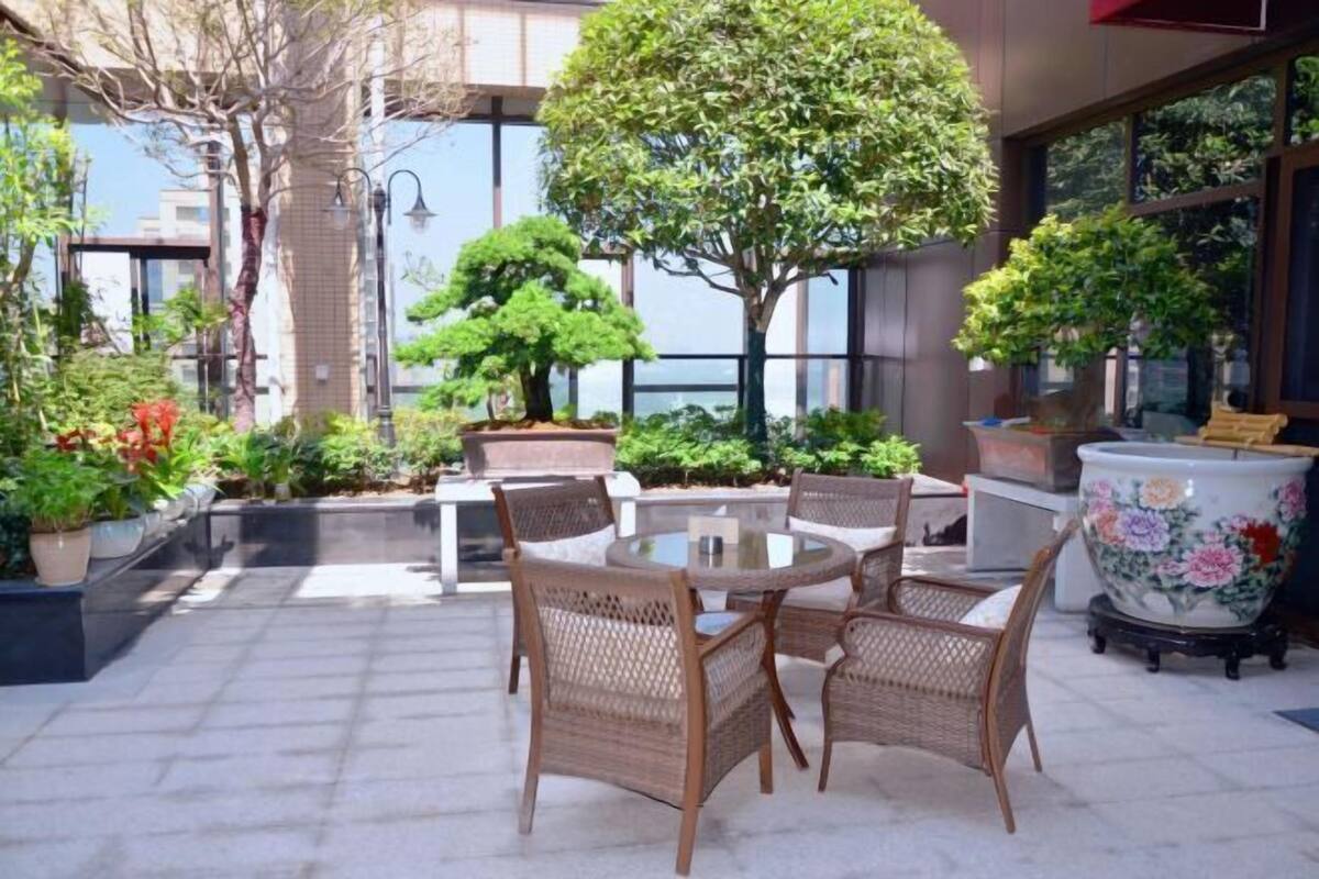 Un espace extérieur aménagé avec des chaises en osier et une table ronde, entouré de plantes en pot et d’un grand arbre.