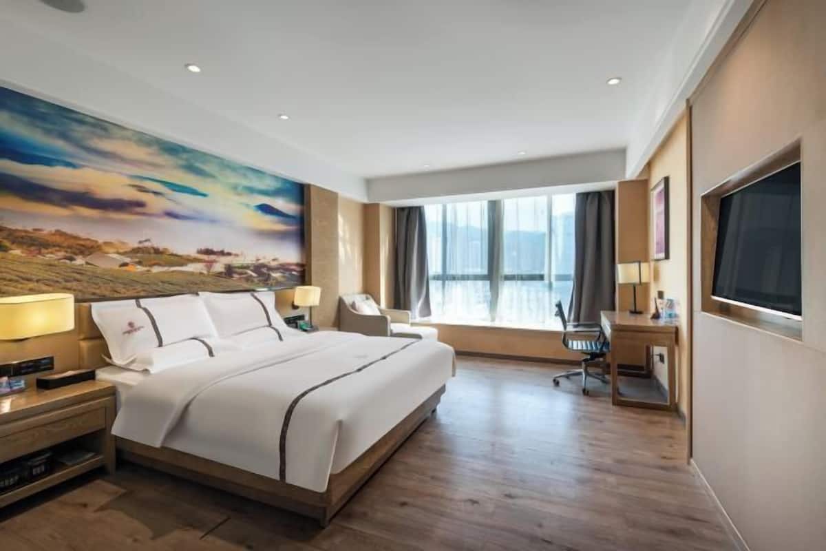 A hotel room with a large bed, a desk, a chair, a TV, and a large window with curtains.