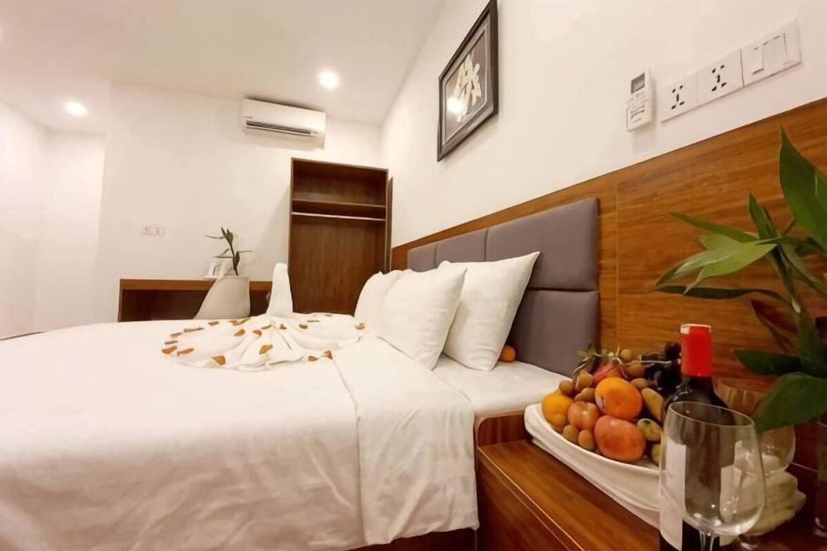 A neatly made bed with a white bedspread, a bedside table with a fruit bowl and a wine glass, and a wooden headboard.