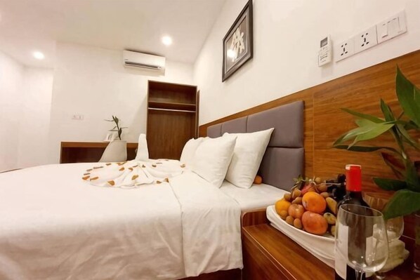 Una cama bien hecha con una colcha blanca, una mesita de noche con un frutero y una copa de vino, y un cabecero de madera.