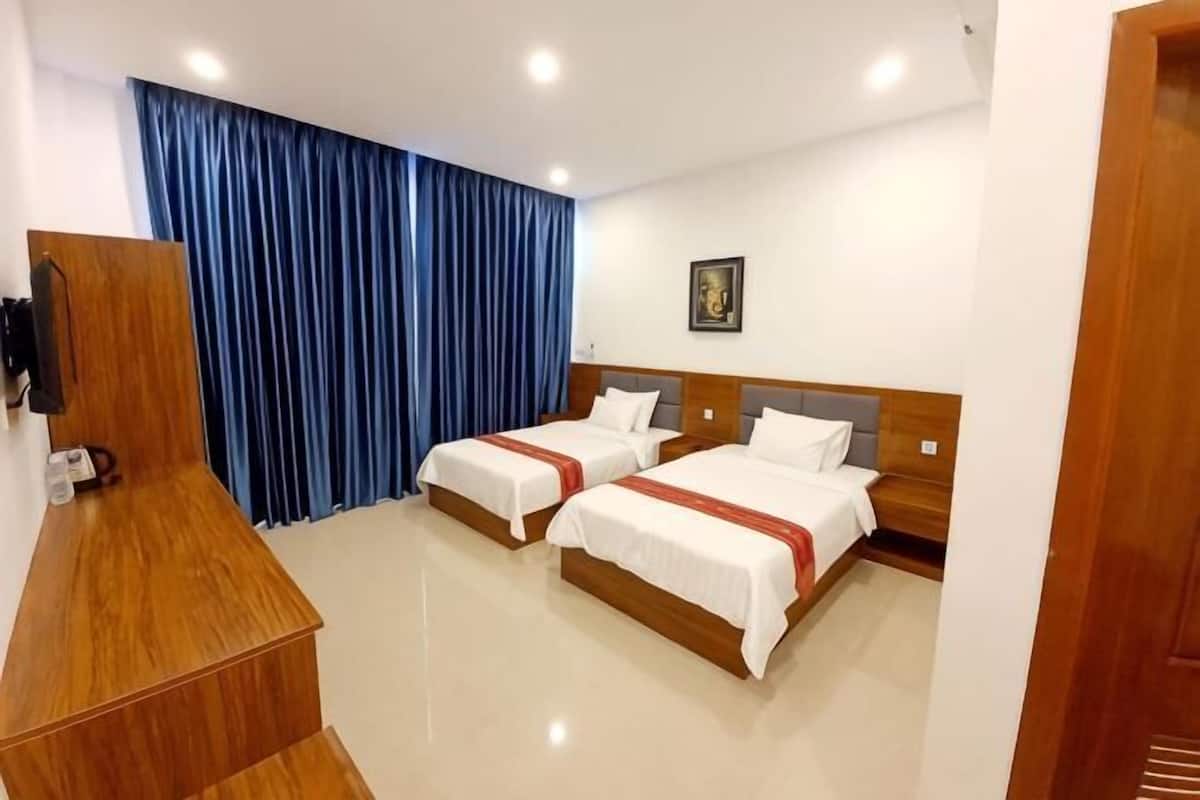 A hotel room with two beds, a wooden desk, a blue curtain, and a framed picture on the wall.