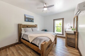 A bedroom with a wooden bed, a ceiling fan, a window with a view, and a small bedside table with a plant. - Oasis at Barkley - Oasis at Barkley (Cadiz)