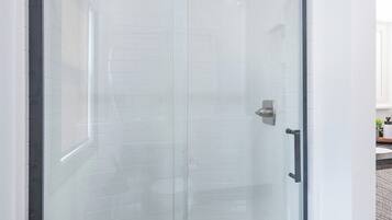 Ducha con cerramiento de vidrio y puerta corrediza, paredes con azulejos blancos y un lavamanos en el mostrador visible.