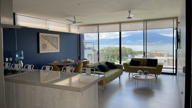 Ein modernes Wohnzimmer mit einem großen Fenster und Blick auf einen See und Berge. Es gibt einen Essbereich mit Tisch und Stühlen sowie eine Couch mit Kissen.