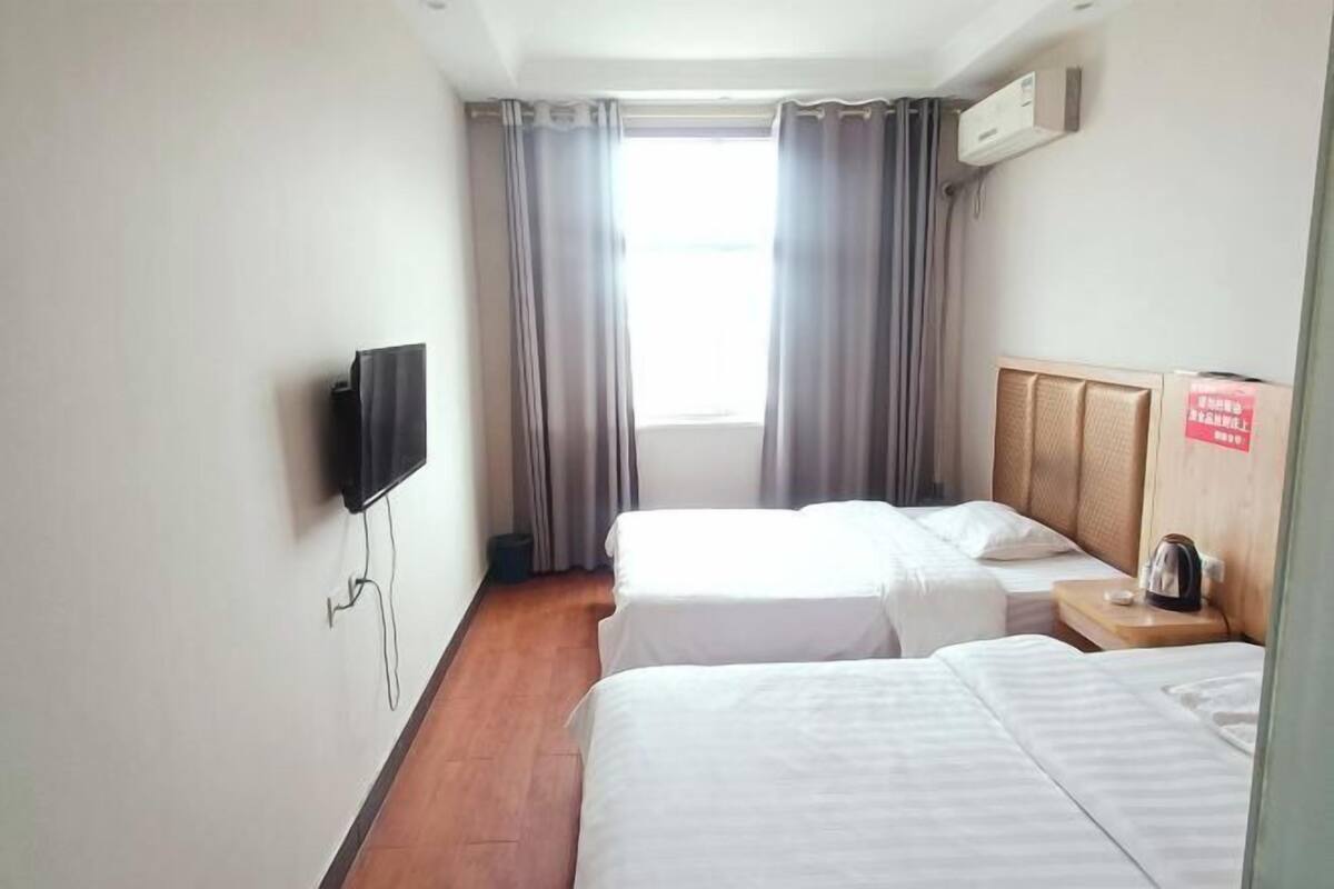 A hotel room with two beds, a TV mounted on the wall, a window with curtains, and a bedside table with a kettle.